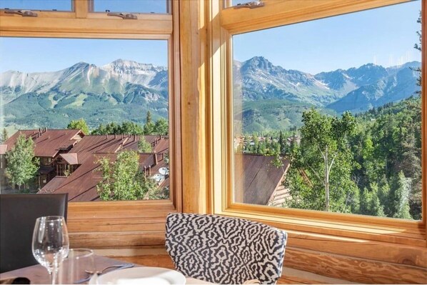 A wooden interior with a table and chairs, a window with a mountain view, and a clear sky.