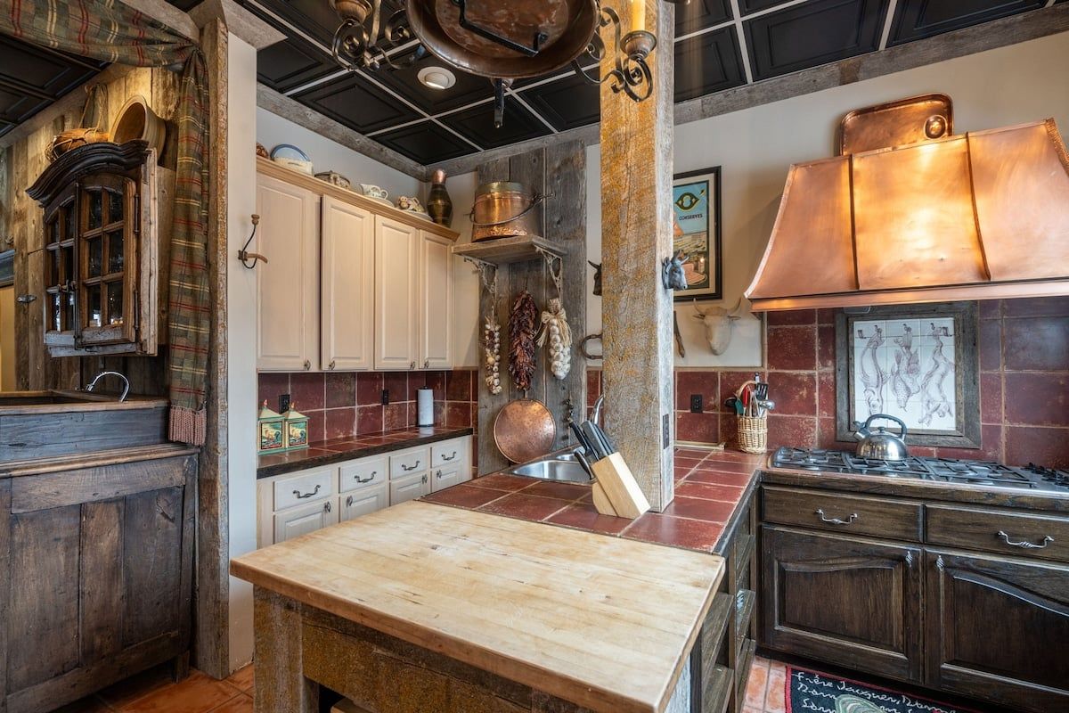 A rustic kitchen with a wooden table, cabinets, and a copper range hood.