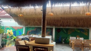 An outdoor seating area with thatched roofs, wicker furniture, and a mural of tropical leaves. - Noah’s Ark Cannabis Bar Restaurant Hostel (Koh Samui)