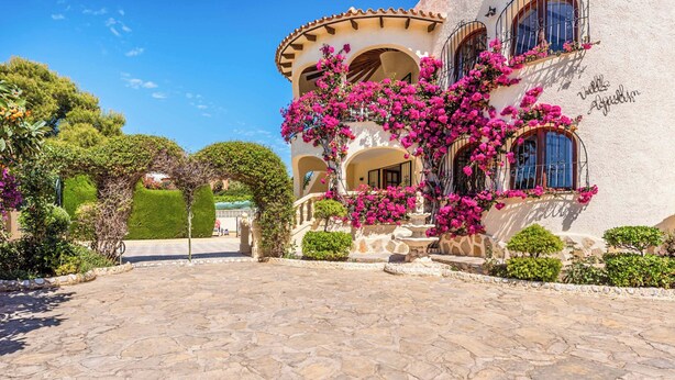 Un edificio con ingresso in pietra, ed edera fiorita di colore rosa, su uno sfondo di cielo azzurro.