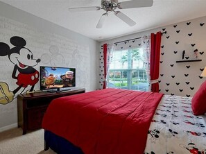 Une chambre avec une grande fresque représentant Mickey Mouse, un lit avec une literie rouge, un téléviseur posé sur un meuble en bois et une fenêtre avec des rideaux rouges.
