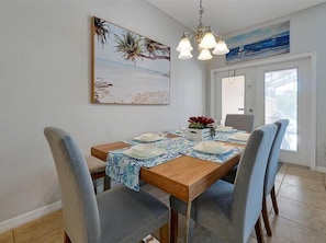 Un coin repas avec une table en bois dressée pour quatre personnes, entourée de chaises. Un tableau représentant une scène de plage orne le mur, un lustre se trouve au-dessus de la table et une fenêtre laisse entrer la lumière naturelle.