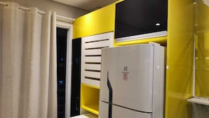 Une cuisine de taille réduite avec un réfrigérateur blanc, un mur d’accent jaune et une fenêtre avec des stores.