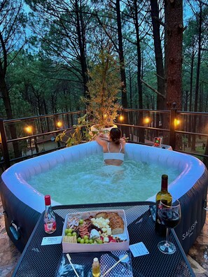 Un jacuzzi rodeado por un bosque, con una mesa en primer plano que sostiene una botella de vino, una copa y una tabla de fiambres.