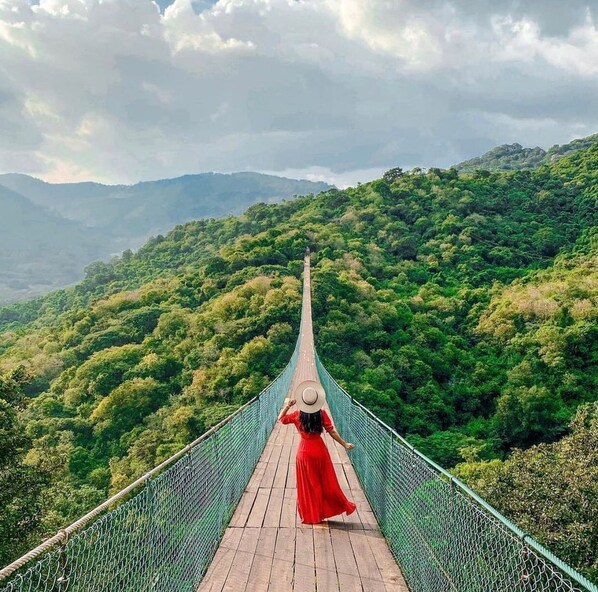 A person in a red dress walking on a suspension bridge with a view of a forested mountain range.