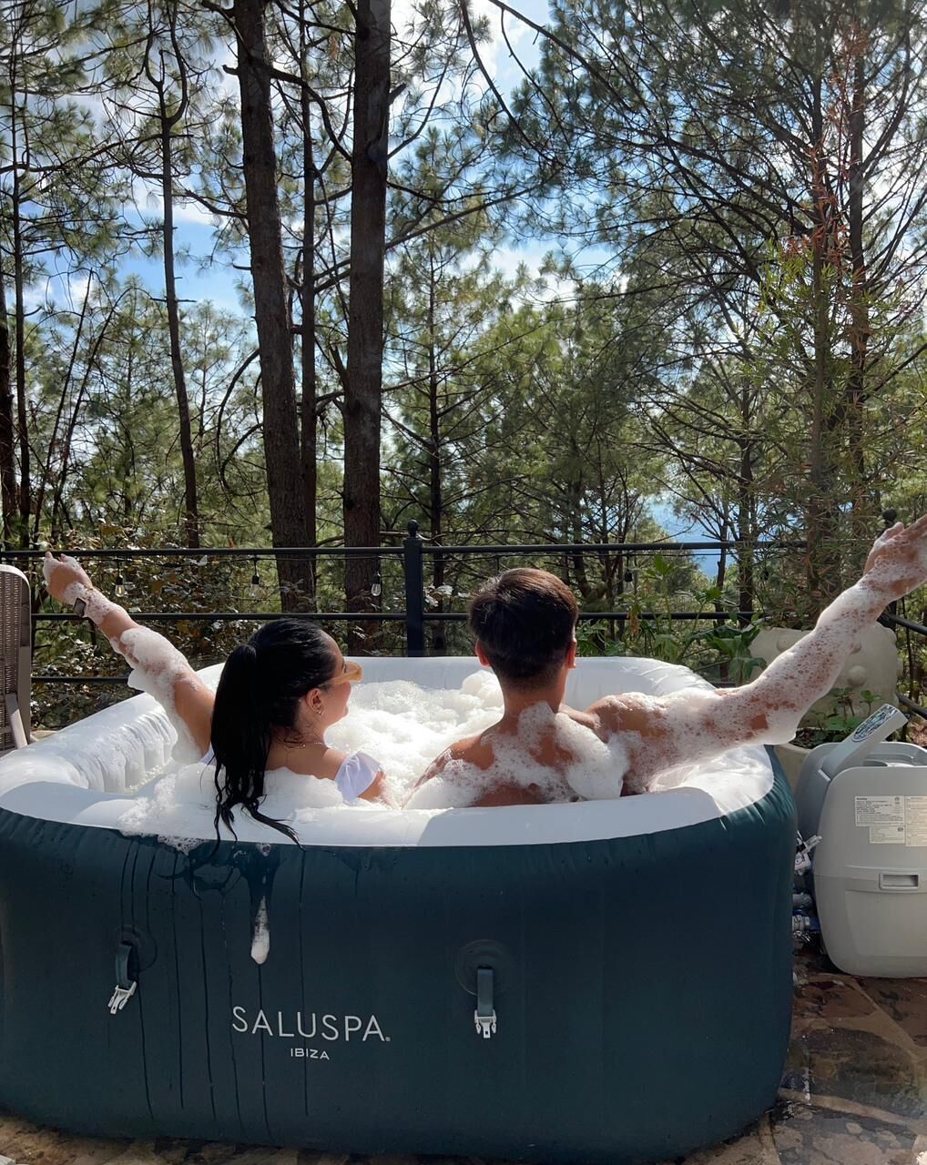 Two people relaxing in a large inflatable hot tub outdoors surrounded by trees.