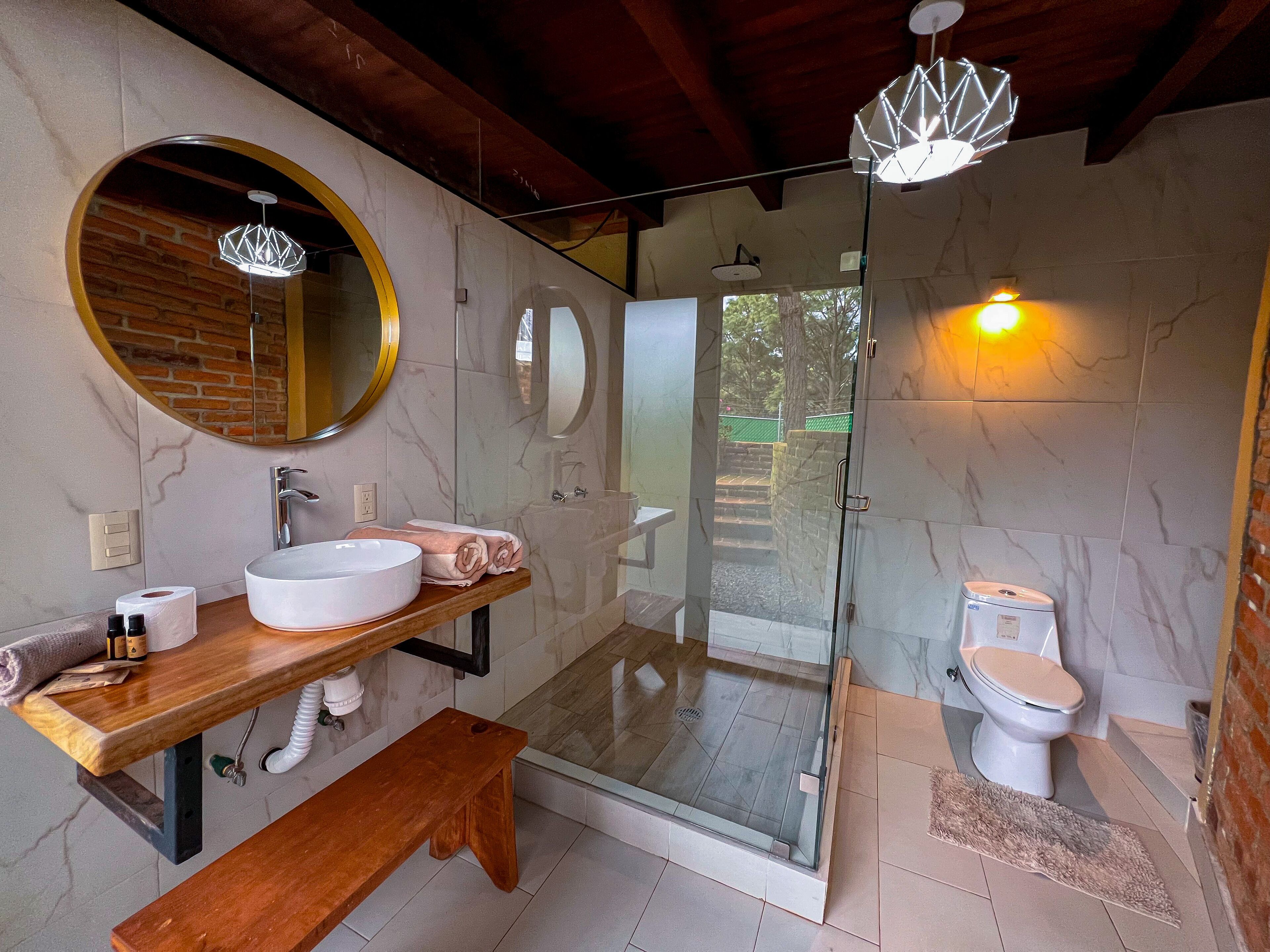 Un baño moderno con tocador de madera, un espejo redondo, ducha con cerramiento de vidrio y un inodoro.