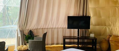 A tent-style room with a bed, a TV, wicker furniture, and a view of greenery outside.