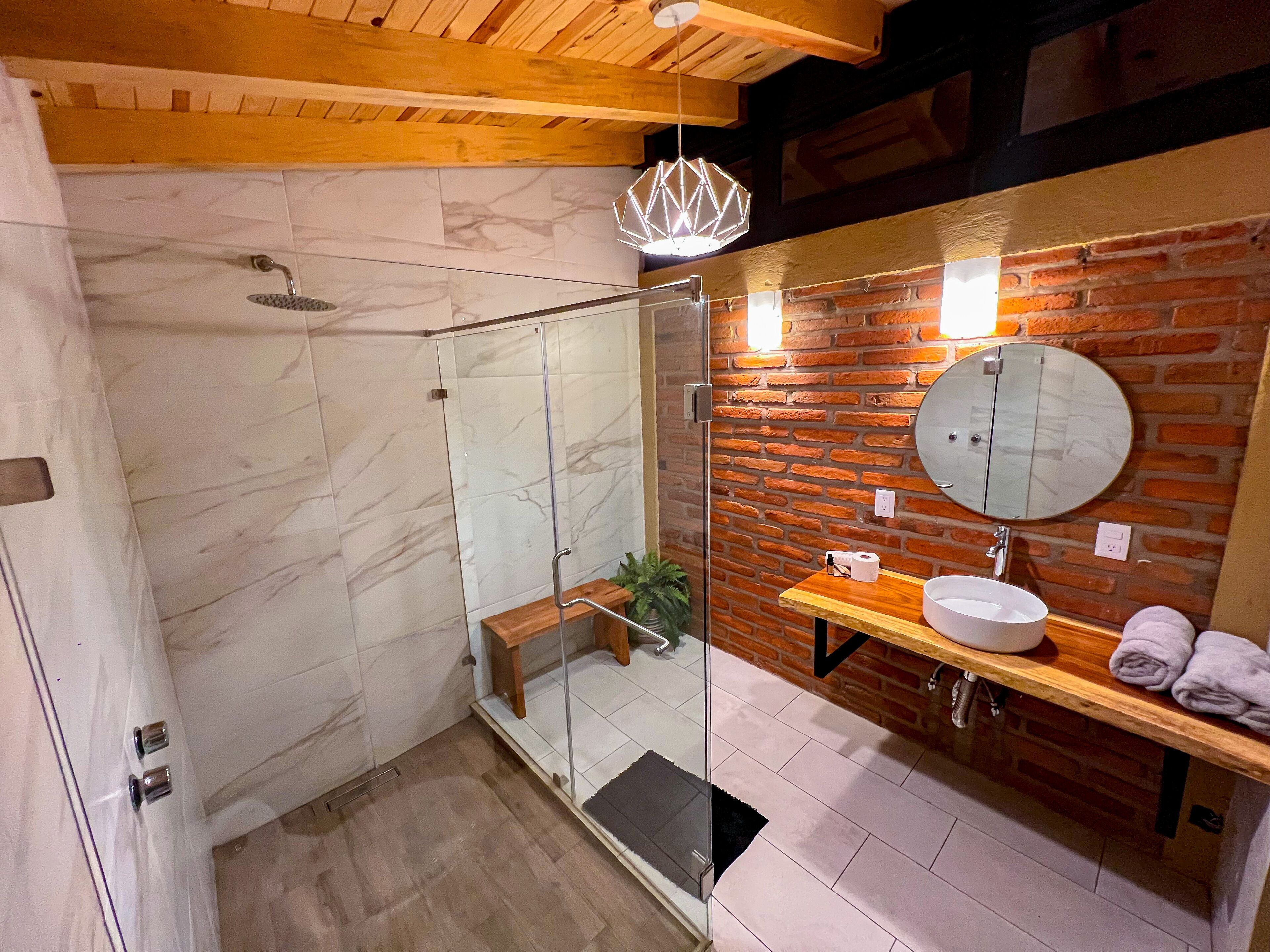 Un baño moderno con ducha envuelta en vidrio, un banco de madera, un espejo redondo y un estante de madera con toallas.