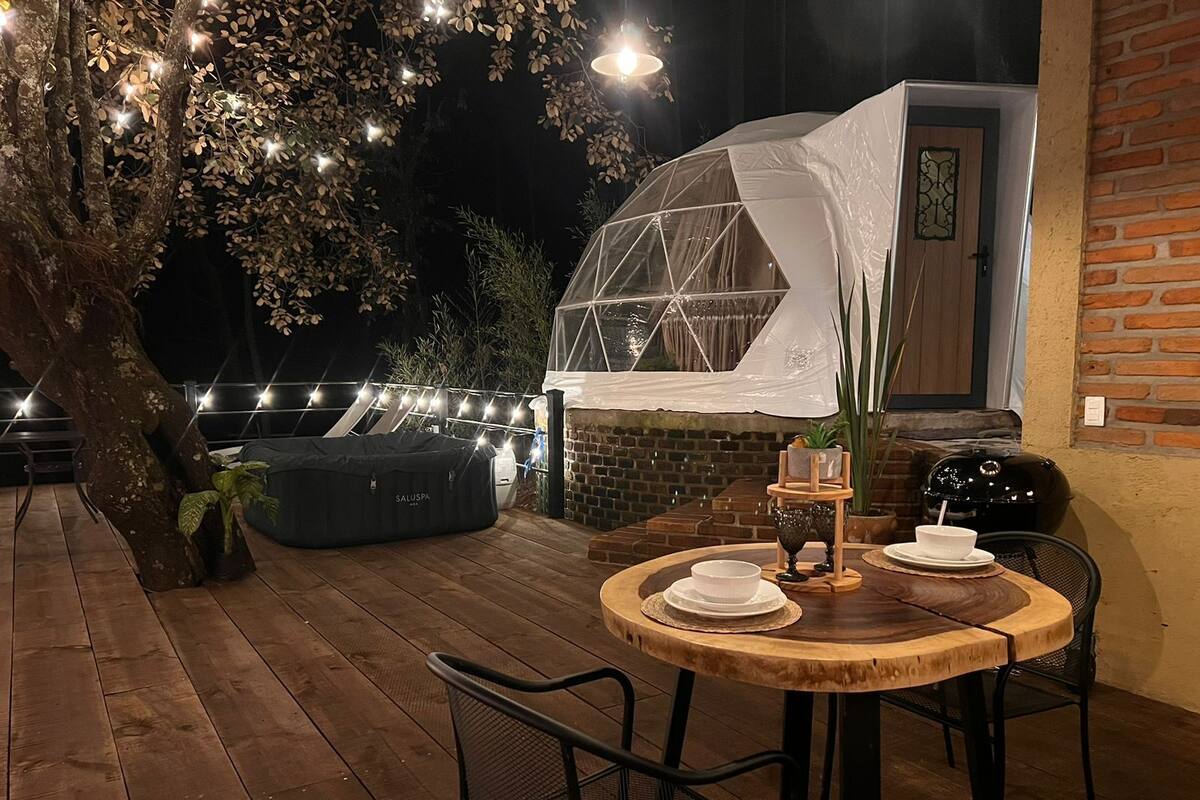 Une terrasse en bois avec une table ronde et des chaises, un espace salon couvert et des guirlandes lumineuses.
