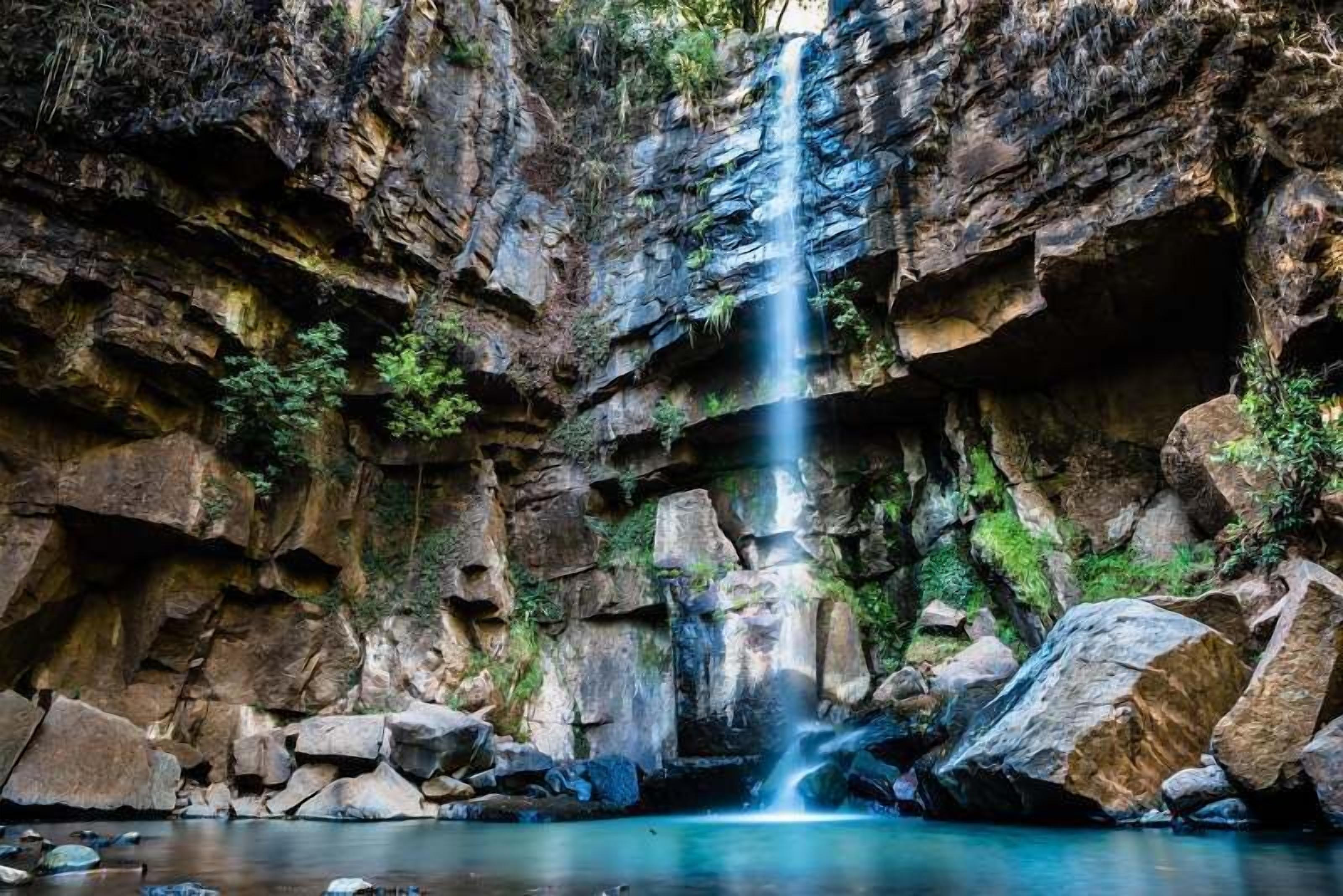Uma cachoeira despencando de um penhasco rochoso em direção a um lago cercado por grandes pedras.