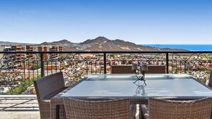 Un balcon doté de meubles en osier et d’une table en verre, offrant une vue sur une ville côtière et les montagnes.