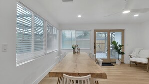 Un vaste espace de salle à manger avec une table en bois et des chaises blanches, de grandes fenêtres équipées de stores et une porte-fenêtre coulissante donnant sur un espace extérieur.