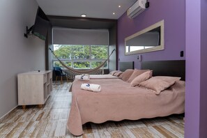A bedroom with a large bed, a desk, a chair, a hammock, and a window with a view of trees. - Hotel El Mesón  (La Dorada)