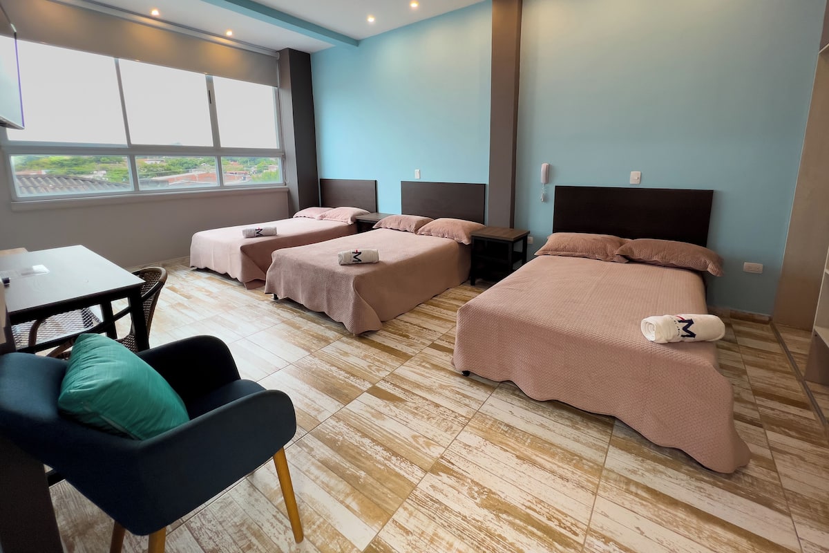 A hotel room with two single beds, a double bed, a small table, a chair, and a view of the outside through large windows.