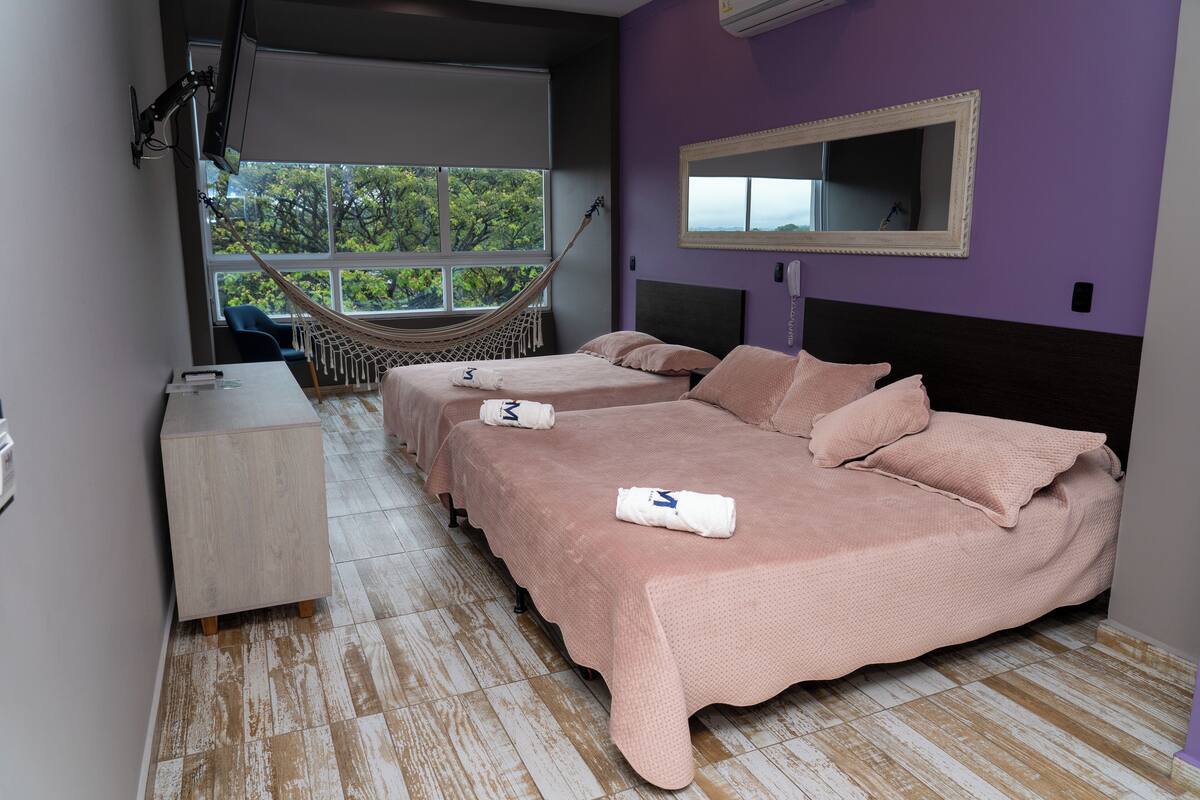 A bedroom with two beds, a hammock, a desk, and a window with a view of trees.