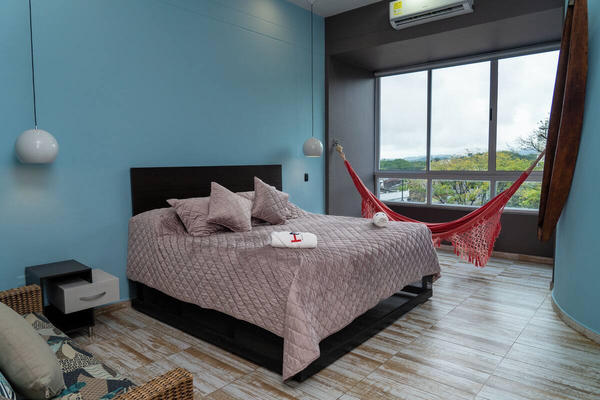 A bedroom with a bed, a nightstand, a hammock, and a large window with a view.