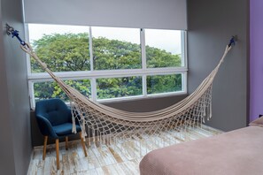 Een hangstoel bij een raam met uitzicht op bomen, een blauwe fauteuil en een bed met een roze deken.