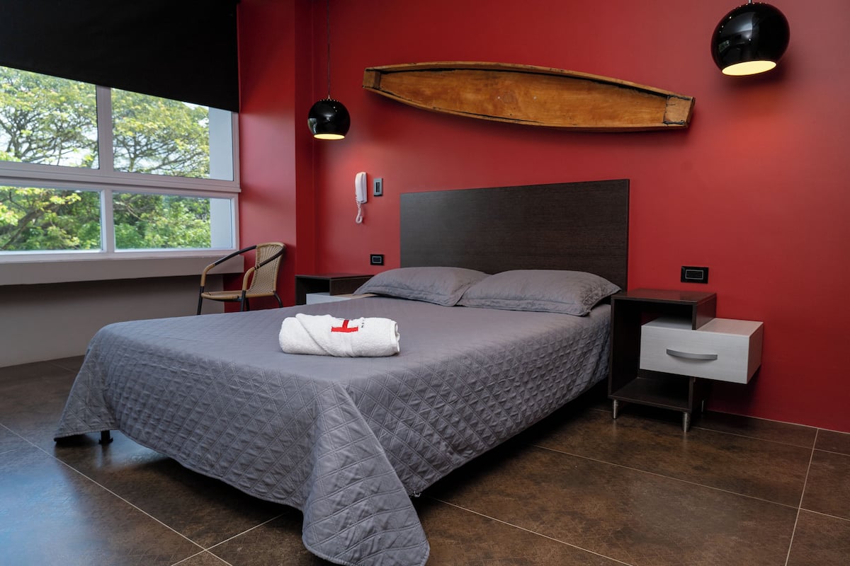 A modern bedroom with a large bed, a wooden boat decoration on the wall, a window with a view of trees, and a chair.