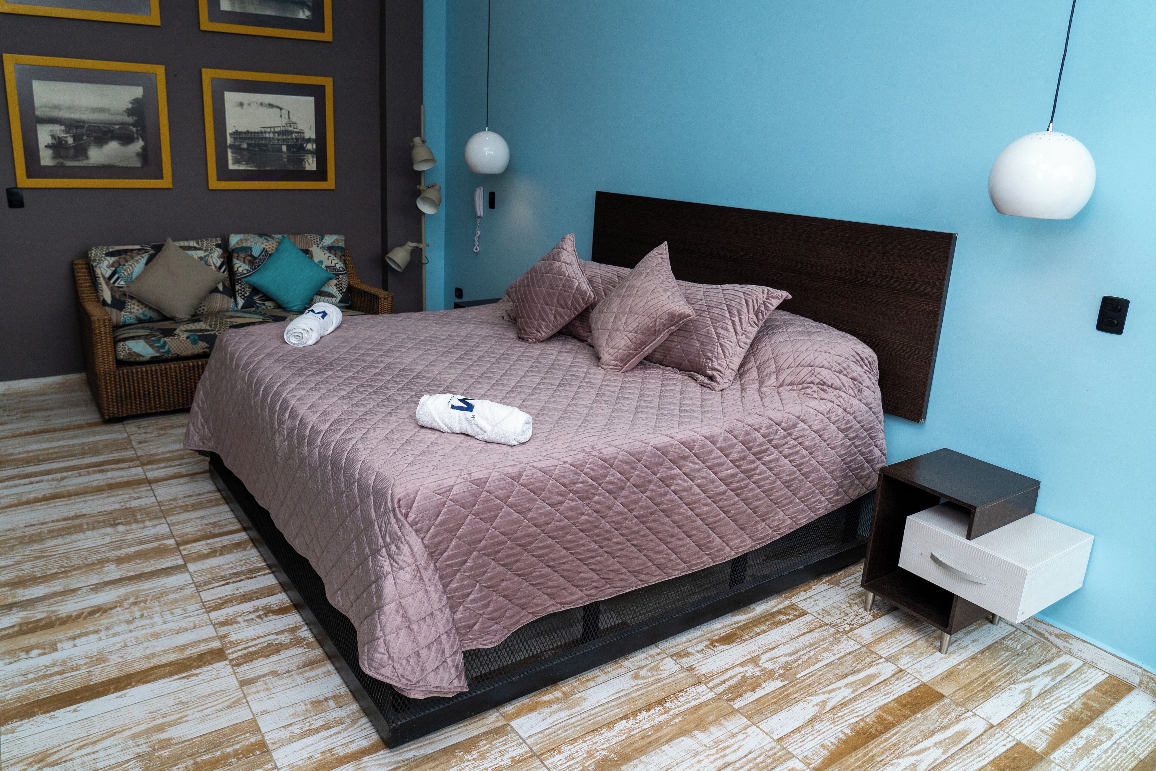 A neatly made bed with a pink quilt, a small bedside table with a drawer, and a wicker chair with cushions.
