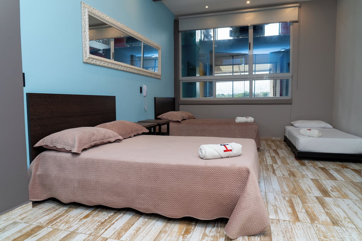 A hotel room with a large bed, a smaller bed, a desk, a mirror, and a window with a view of the outside.