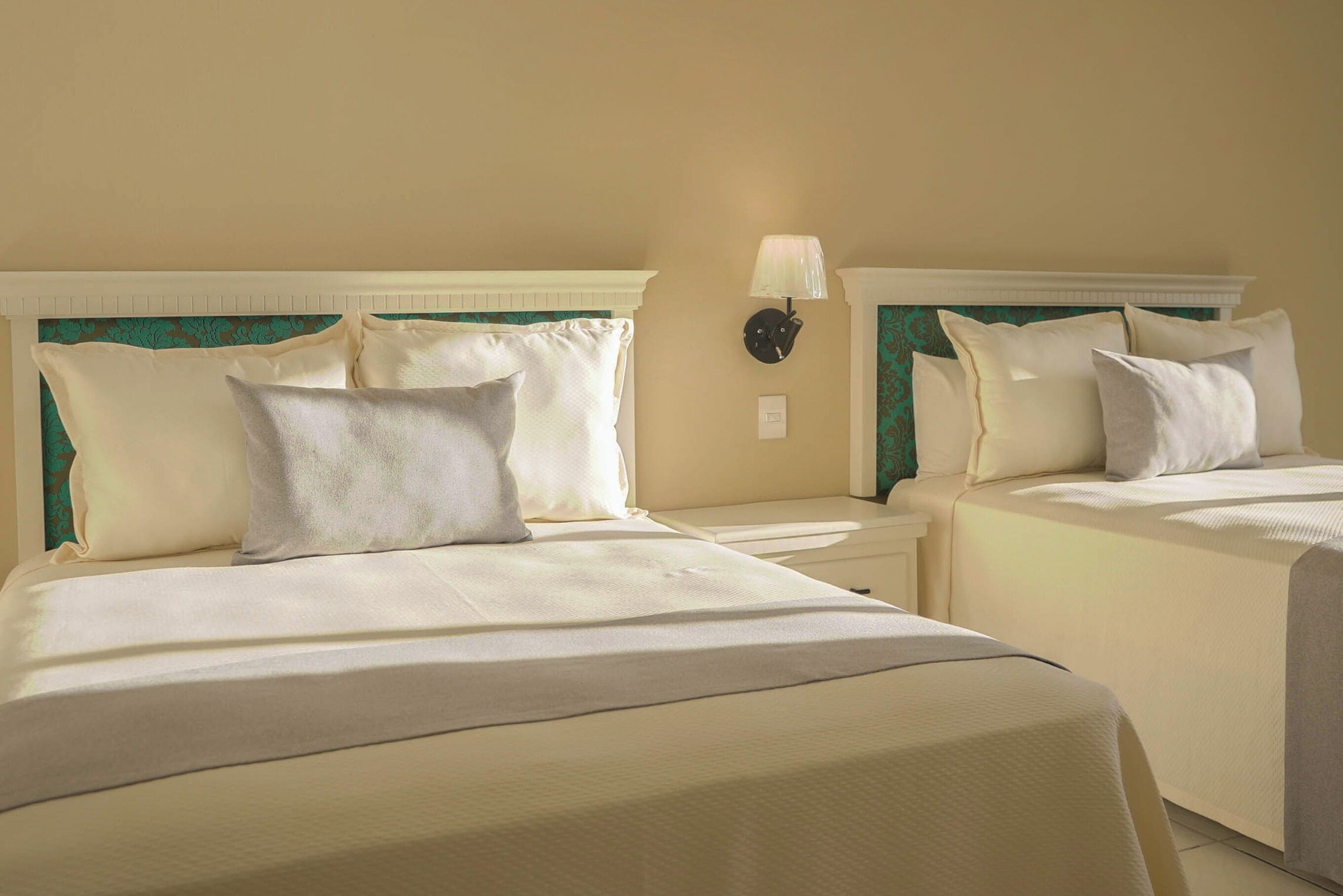 A hotel room with two beds, each with a white bedspread and multiple pillows. There is a wall-mounted lamp on the wall between the beds.