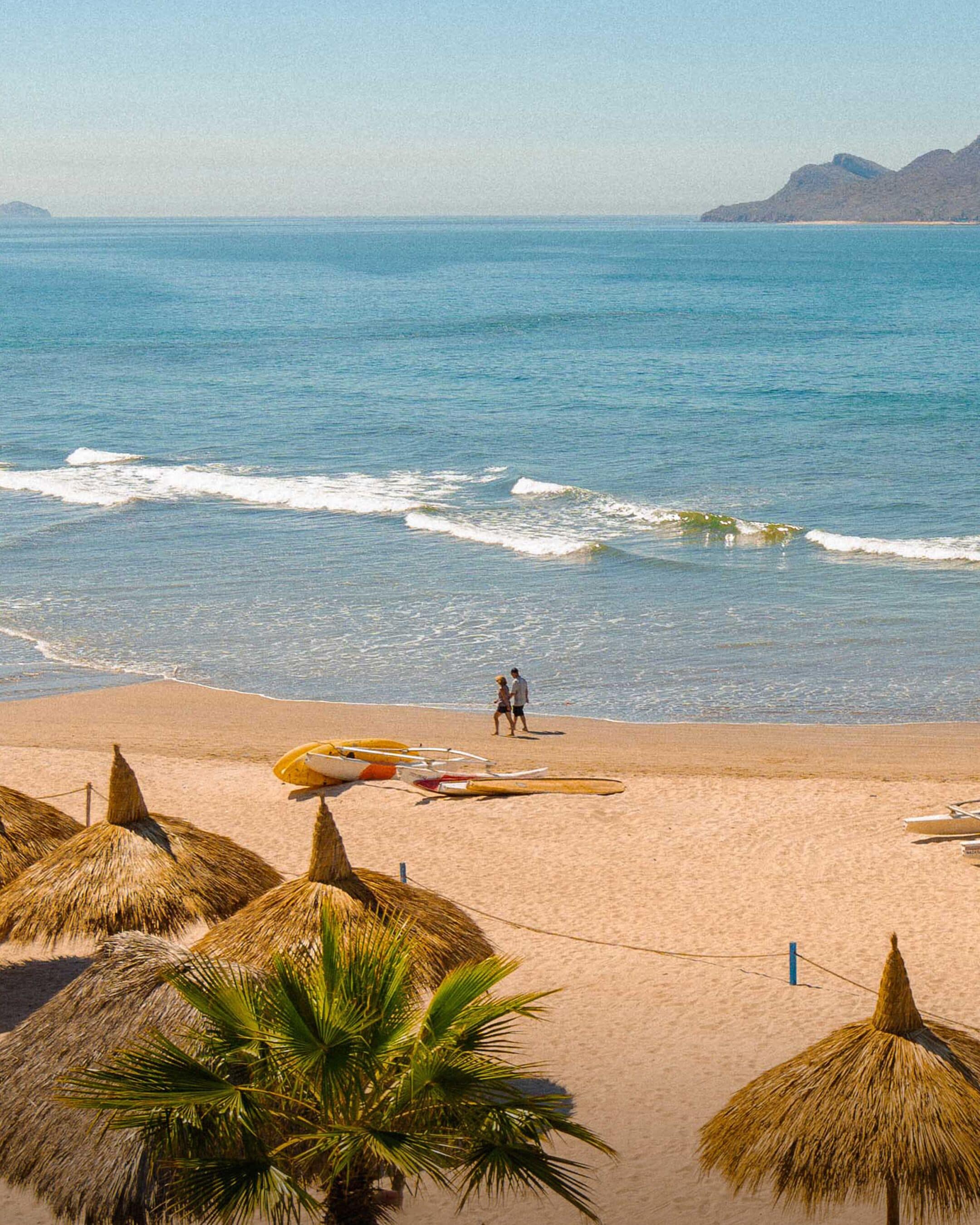 A beach with thatched umbrellas, a couple walking, and surfboards on the sand.