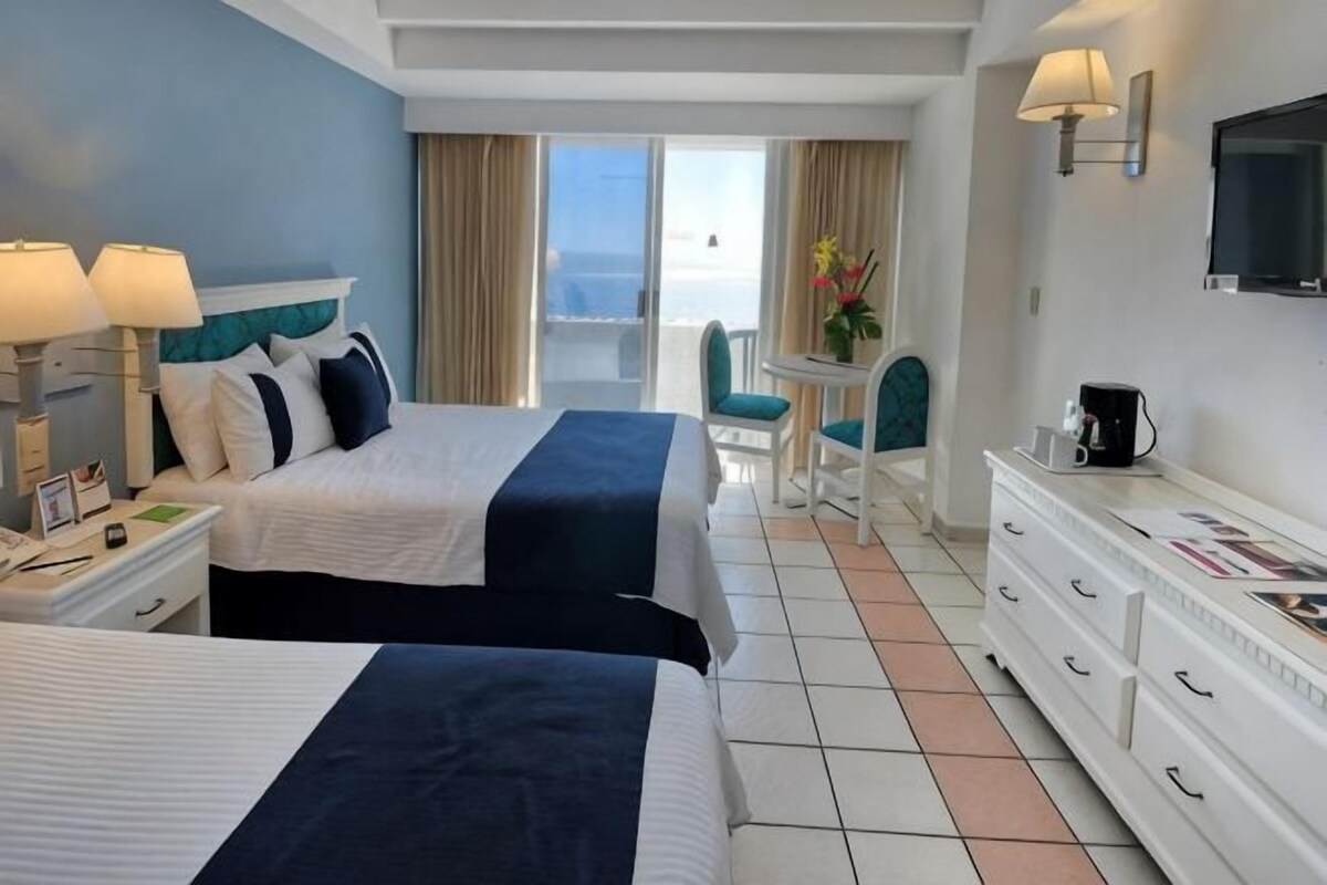 A hotel room with two beds, a TV, a desk with a chair, and a view of the outdoors.