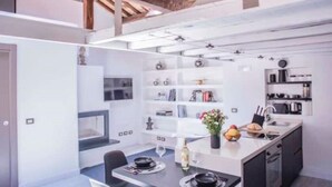 A modern kitchen with white cabinetry, a central island with a sink, and a dining area with a table and chairs. - Mad Hatter (Roma)