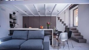 A modern living space with a grey sofa, a dining area with a table and chairs, a kitchen area with cabinets, and a staircase leading to an upper level. - Mad Hatter (Roma)
