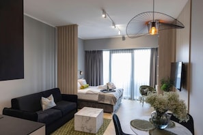 A modern hotel room with a bed, a sofa, a dining table, and a TV.