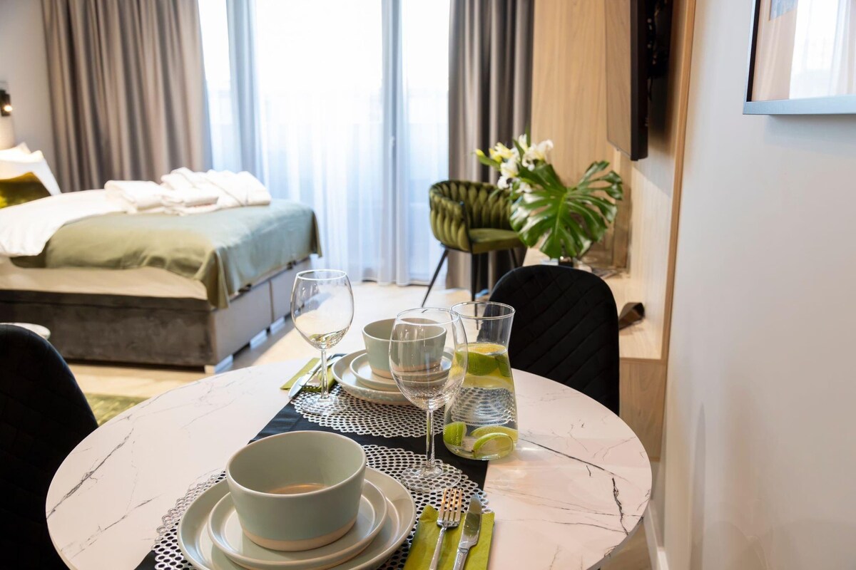Une chambre d’hôtel avec un lit soigneusement fait, une table à manger dressée avec des assiettes, des verres et des couverts, et une grande fenêtre avec des rideaux.