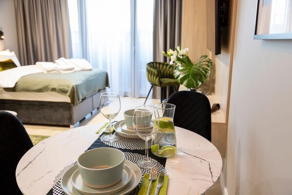 A hotel room with a neatly made bed, a dining table set with plates, glasses, and cutlery, and a large window with curtains.