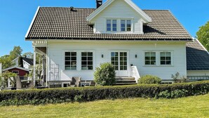 Une maison blanche à deux étages, avec un toit en tuiles, une véranda à l’avant équipée de mobilier d’extérieur et une pelouse bien entretenue.