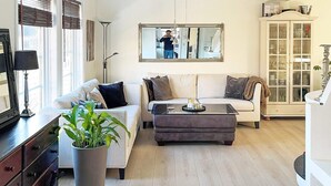 Un salon comprenant un canapé, un pouf et une table basse. On y trouve également une plante en pot, une lampe et un meuble avec des portes en verre.