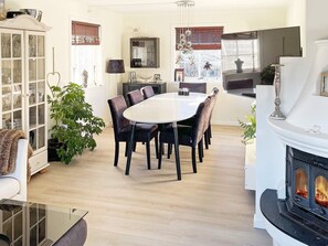 Ein modernes Wohnzimmer mit Kamin, einem Essbereich mit rundem Tisch und Stühlen, einer Vitrine mit Dekorationen und einem großen Fenster mit Jalousien.