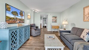 A living room with a blue cabinet, grey sofa, and a coffee table. There is a painting of palm trees on the wall and a wooden sign on the cabinet. - Bluewater Resort 207 (Myrtle Beach)