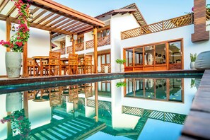 Un espace piscine agrémenté de chaises longues et de tables en bois, une piscine aux eaux cristallines qui reflète l’environnement, et un bâtiment à deux étages doté de grandes fenêtres et de balcons en bois.