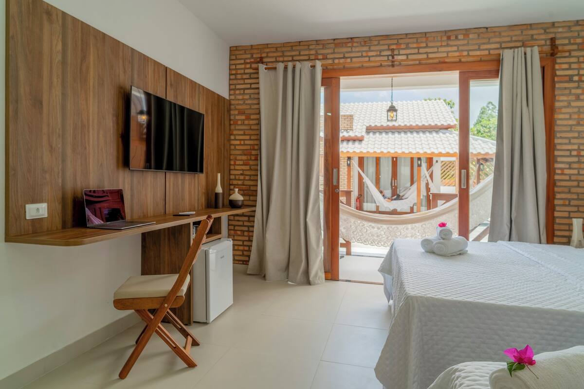 Une chambre d’hôtel avec un téléviseur fixé au mur, un bureau avec un ordinateur portable, un petit réfrigérateur, un lit recouvert d’une courtepointe blanche et un hamac à l’extérieur de la porte.