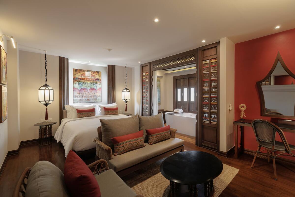 Quarto de hotel com cama, sofá, mesa de jantar e uma cadeira. Há uma janela com cortinas, televisão na parede e um espelho decorativo.