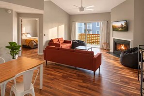 A modern living room with a wooden floor, a sofa, a dining table with chairs, a fireplace, and a balcony with a view. - Corporate Retreat: Stylish 2BR, Pool, Balcony (Wilmington)