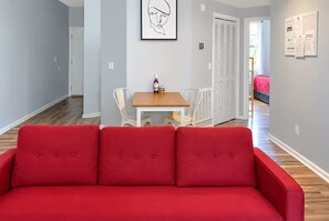 A red sofa in a living area with a dining table and chairs, a white door, and a framed picture on the wall. - Corporate Retreat: Stylish 2BR, Pool, Balcony (Wilmington)