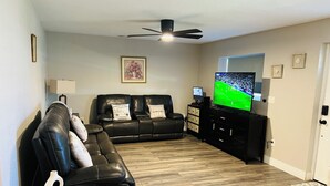 Un salon avec un canapé en cuir noir, un téléviseur à écran plat, un ventilateur de plafond et un plancher en bois.