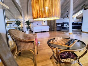 Un vaste espace de vie comprenant une table ronde en verre pour jouer aux échecs, des fauteuils en osier et un luminaire suspendu.