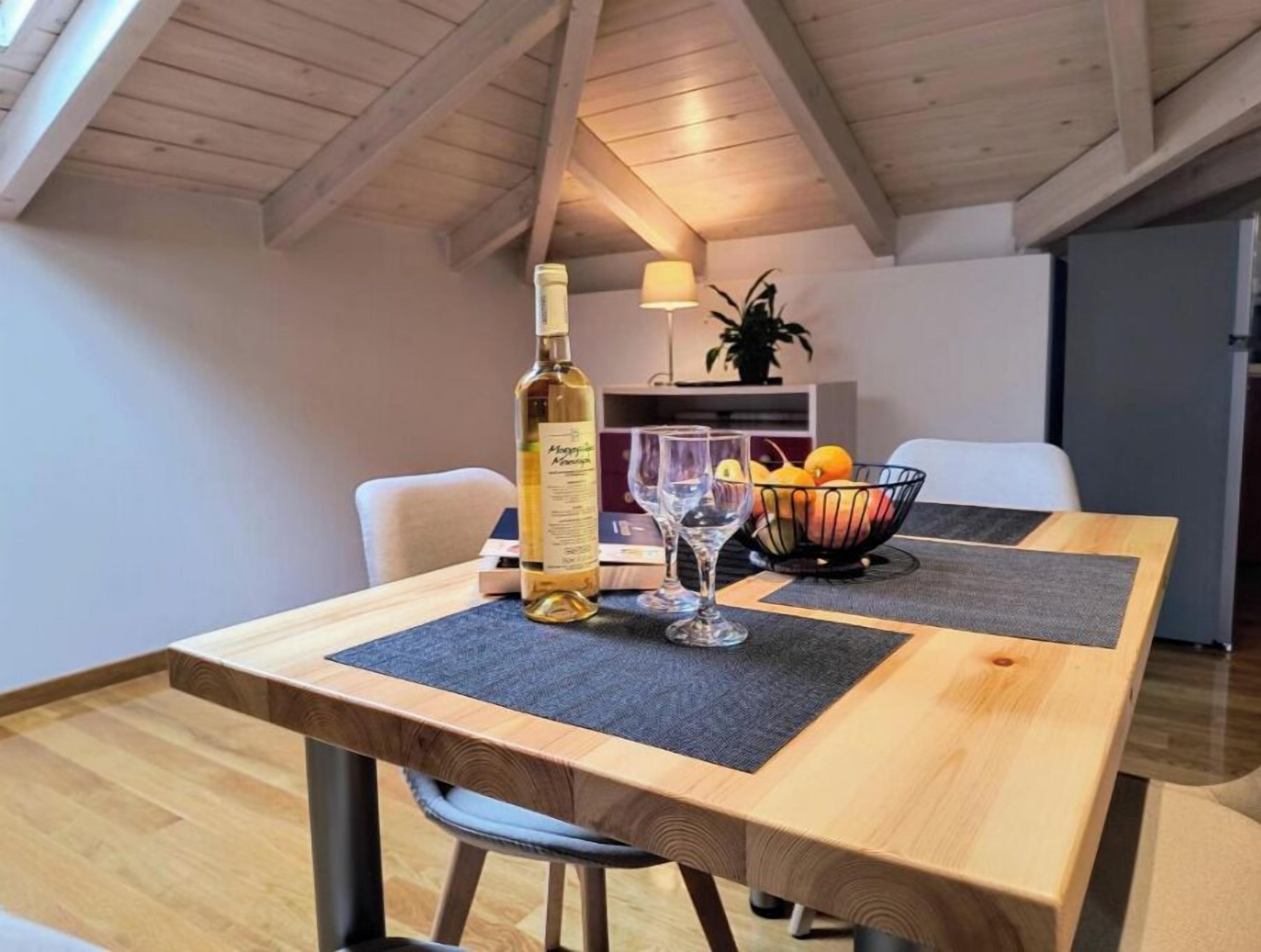 A dining table with a bottle of wine, two glasses, and a fruit bowl in a modern room with wooden beams and a plant.