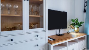 A room with a TV on a wooden stand, a cabinet with glass doors, and a plant on the stand. - Tenerife Royal Gardens - Beachfront Bliss. Studio Apartment Sea/Pool views. (Playa de la Américas)
