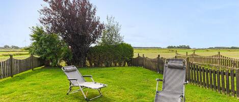 Two lawn chairs in a grassy area with a wooden fence and a tree in the background.