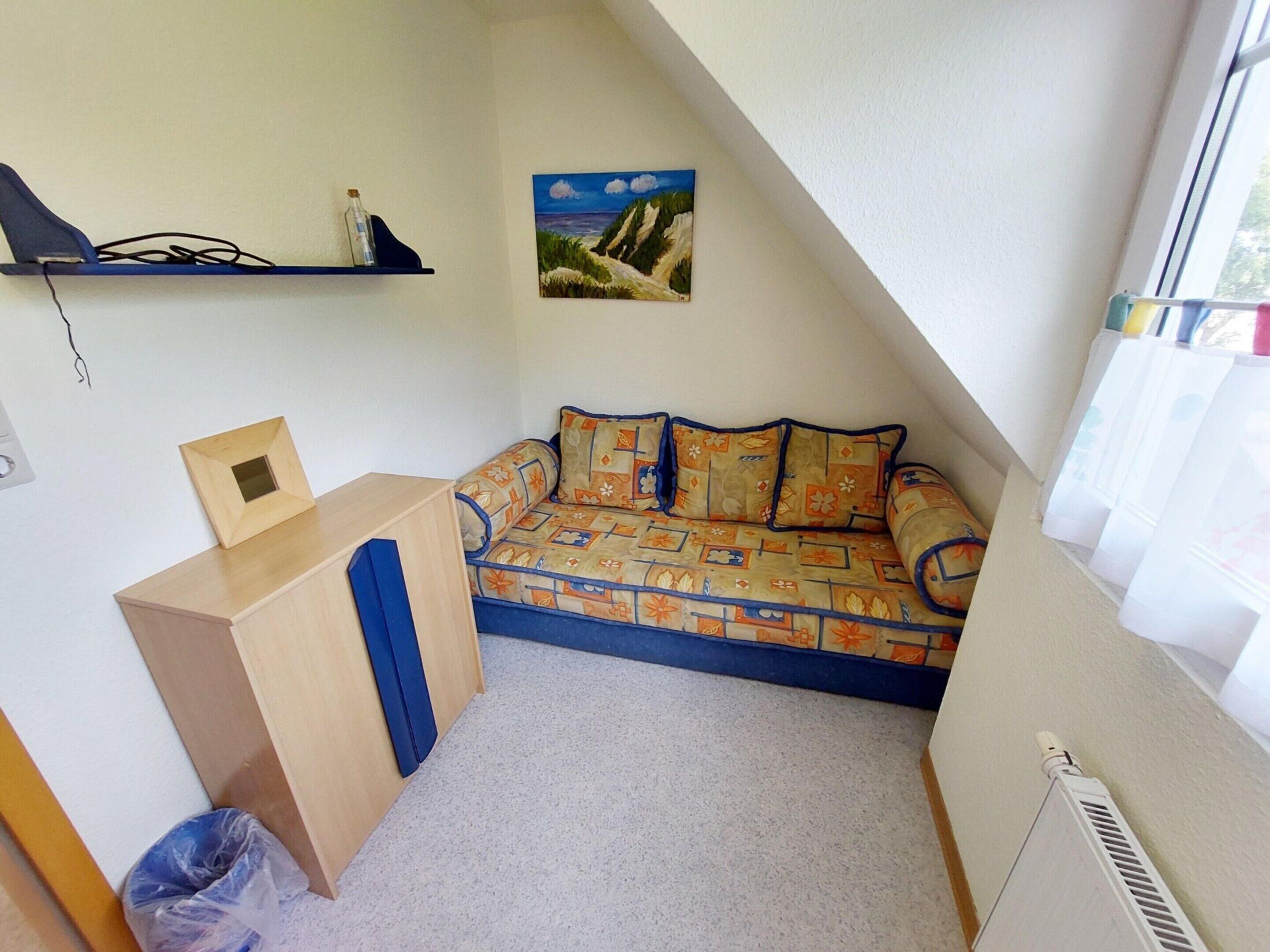 Une petite pièce chaleureuse avec un plafond en pente, un canapé agrémenté de coussins à motifs, un petit bureau en bois, un cadre photo et un tableau accroché au mur.