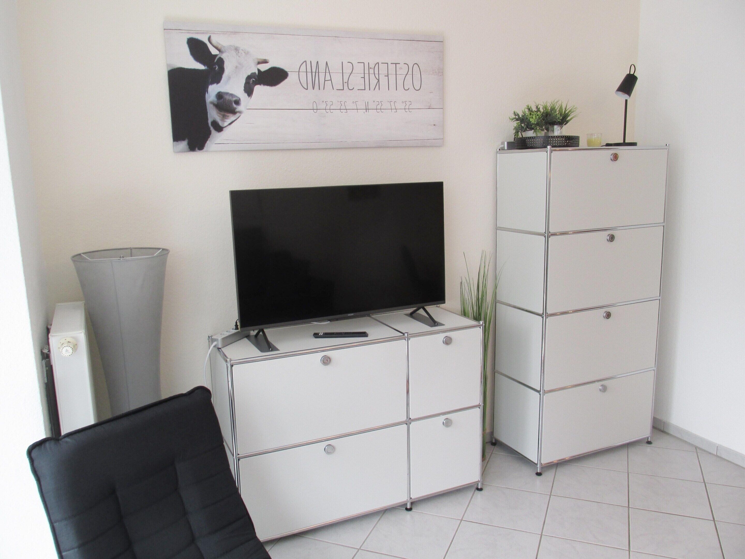 Una sala de estar con un televisor de pantalla plana sobre un mueble blanco, un sillón negro y un adorno en la pared.