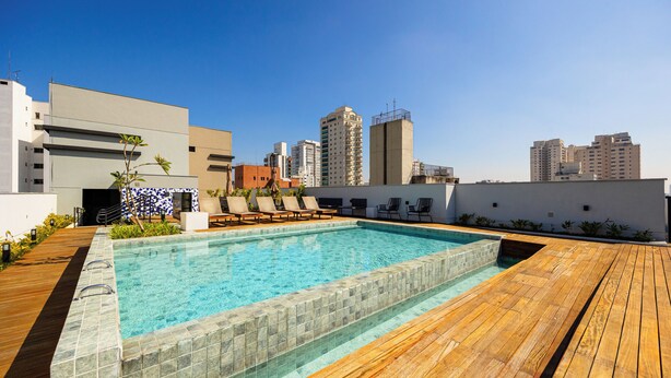 A rooftop pool with a wooden deck, lounge chairs, and a cityscape view. - OCB - Luxo e Conforto Campo Belo (São Paulo)